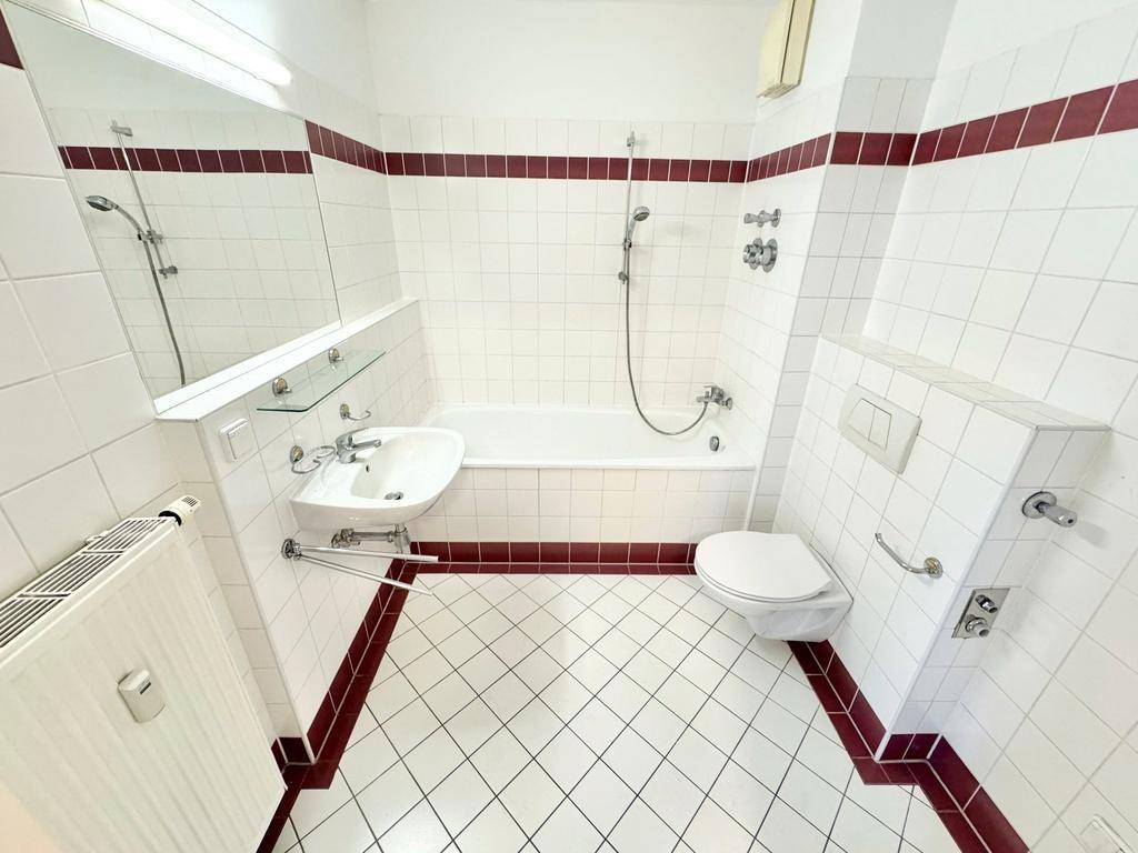 Badezimmer mit weißen Fliesen, rotem Akzentstreifen, Badewanne mit Duschfunktion und WC.