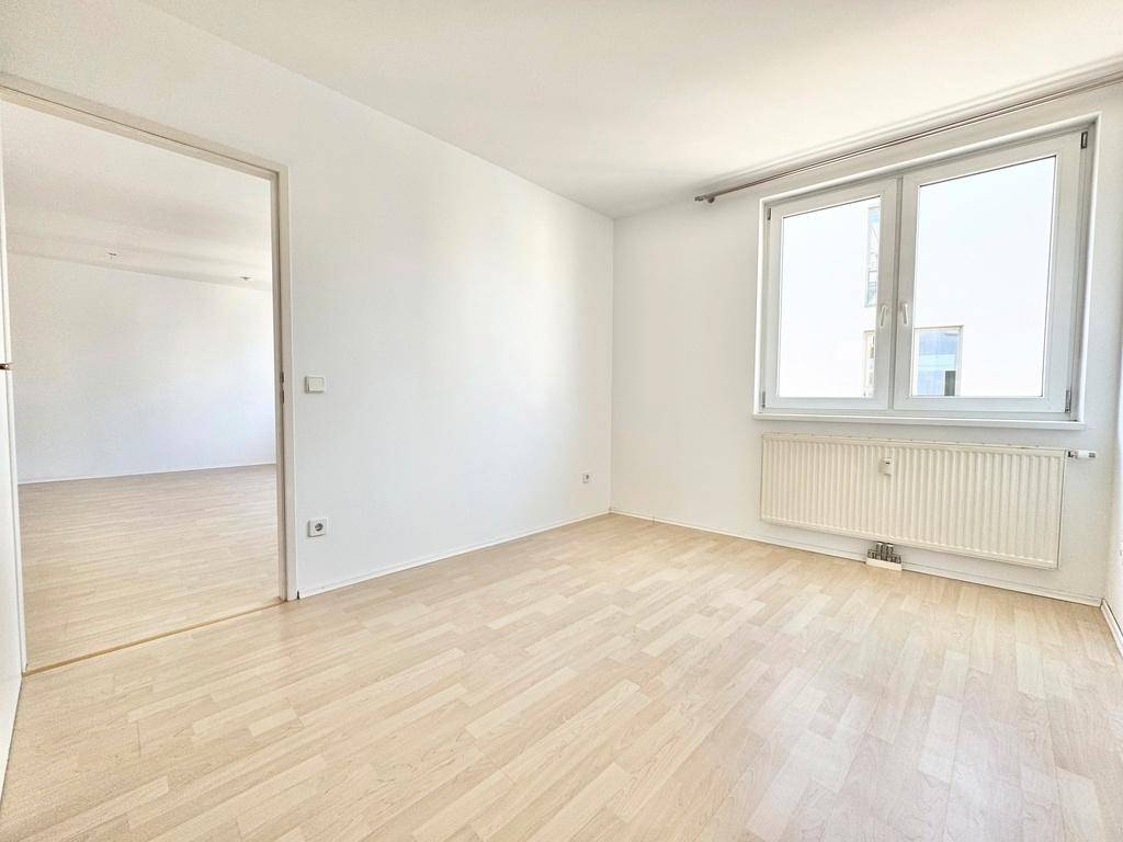 Helles Schlafzimmer mit Parkettboden und großem Fenster, verbunden mit einem weiteren Raum.