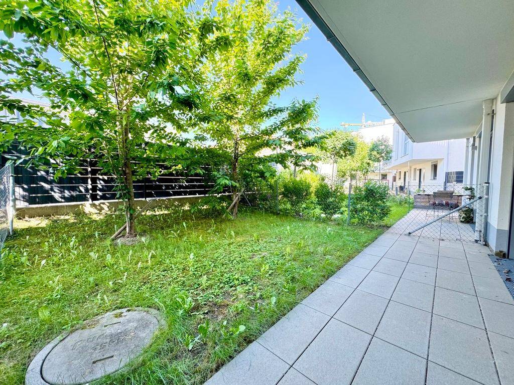 Sonniger Gartenbereich mit gepflasterter Terrasse und jungem Baumbestand.