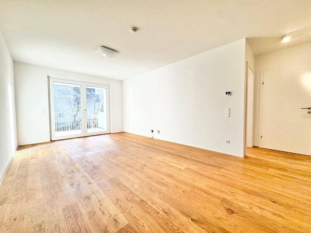 Helles, leeres Wohnzimmer mit neuem Holzparkettboden und großem Balkonzugang.