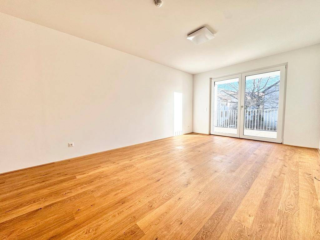 Geräumiges Wohnzimmer mit hellem Holzfußboden und viel Tageslicht durch die breite Balkontür.