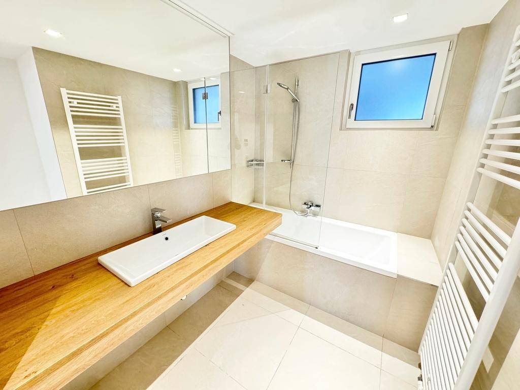 Modernes Badezimmer mit hellen Fliesen, Holzwaschtisch, Badewanne mit Duschfunktion und Fenster.