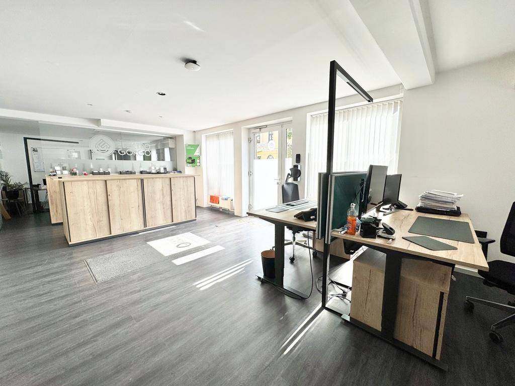 Ansicht des Bürobereichs mit Empfangstheke, Schreibtischen und Computern, durch große Fenster hell erleuchtet.