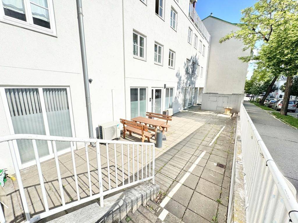 Eine weitere Ansicht der gepflasterten Terrasse mit Holzbänken und Tischen, angrenzend an eine Straße mit Bäumen.