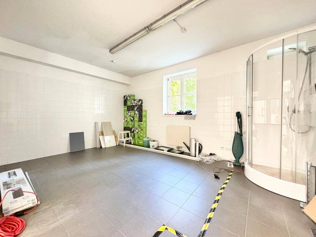 Raum mit weißen Fliesenwänden, dunklem Fliesenboden, einem Fenster und einer Duschkabine, als Hauswirtschaftsraum genutzt.