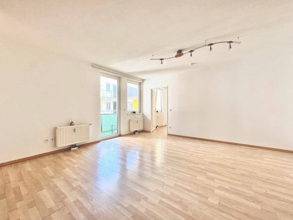 Helles, leeres Wohnzimmer mit Laminatboden, weißen Wänden und Zugang zum Balkon.