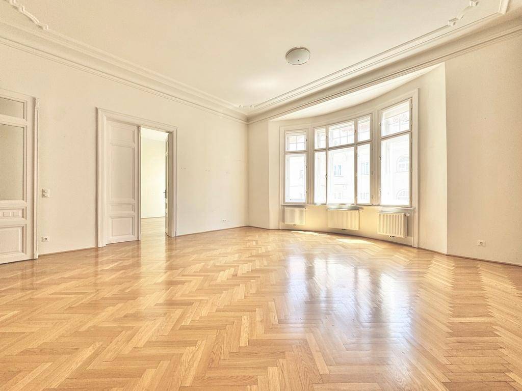 Helles Wohnzimmer mit Fischgrätparkett, hohen Decken und einem großen Erkerfenster, das viel Licht hereinlässt.