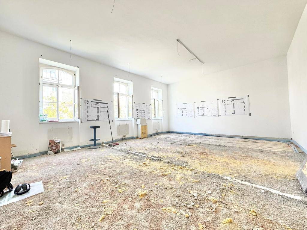 Großer, leerer Raum in Renovierung mit freigelegtem Boden und viel Tageslicht durch hohe Fenster.