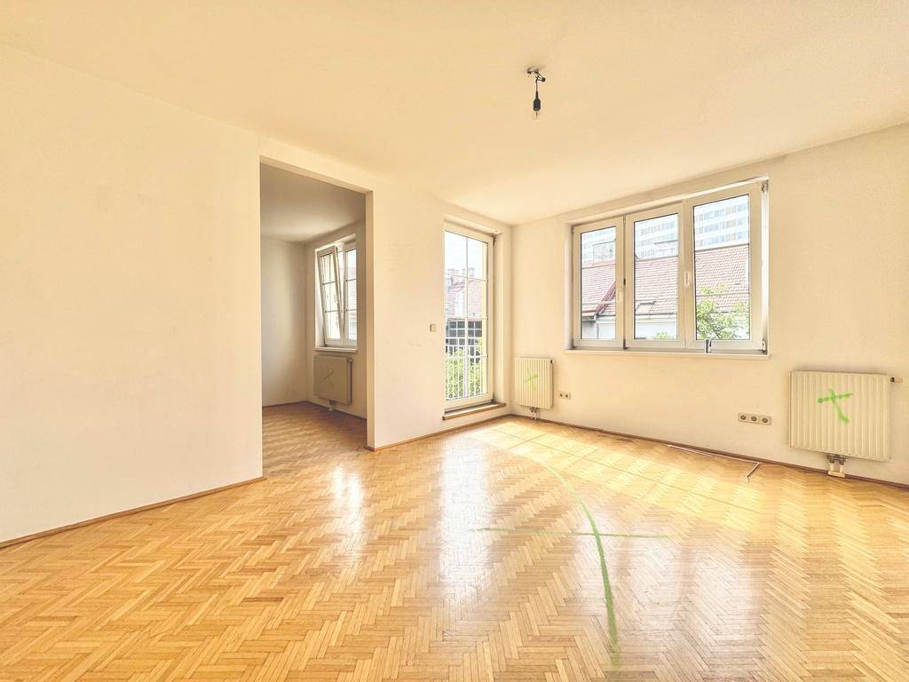 Lichtdurchflutetes Wohnzimmer mit klassischem Parkettboden, weißen Wänden und Zugang zum Balkon.