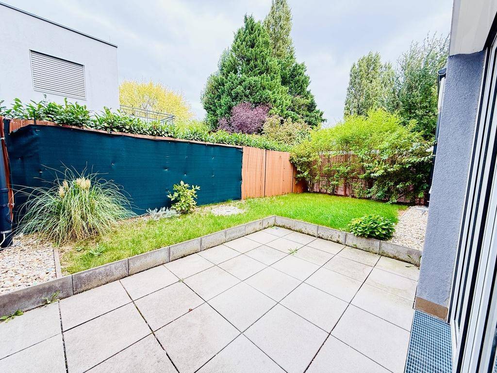 Sonniger Außenbereich mit gepflasterter Terrasse und kleinem Rasenstück, umgeben von Grün.
