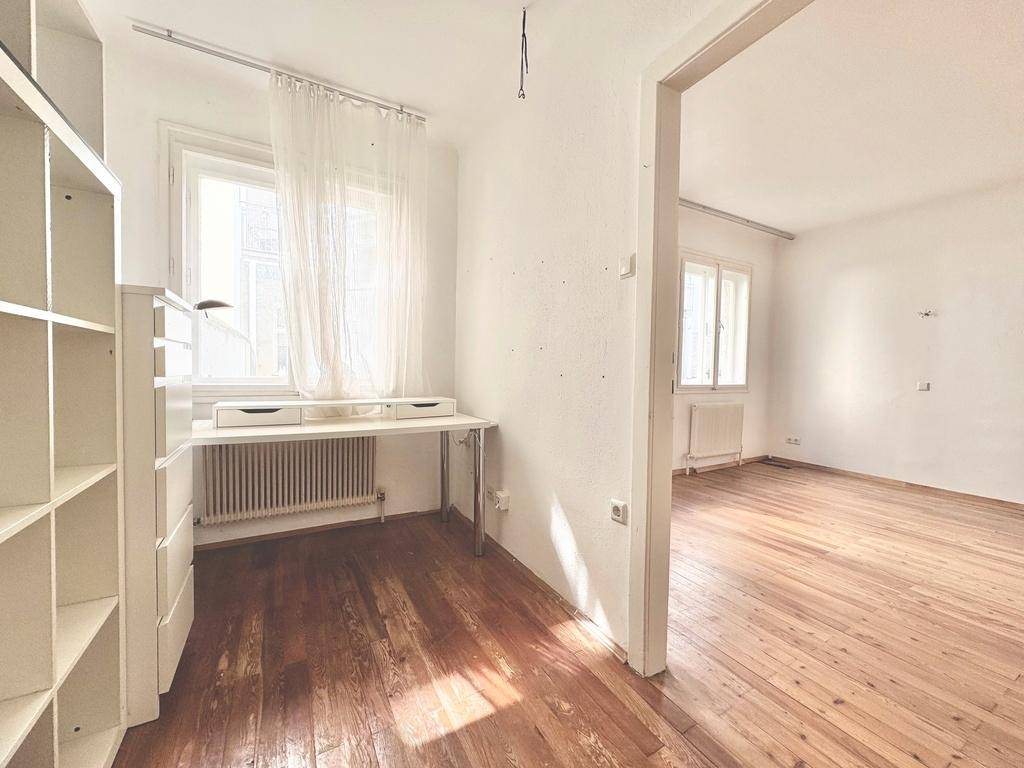 Helles Arbeitszimmer mit Holzboden, Schreibtisch am Fenster und einem Bücherregal.