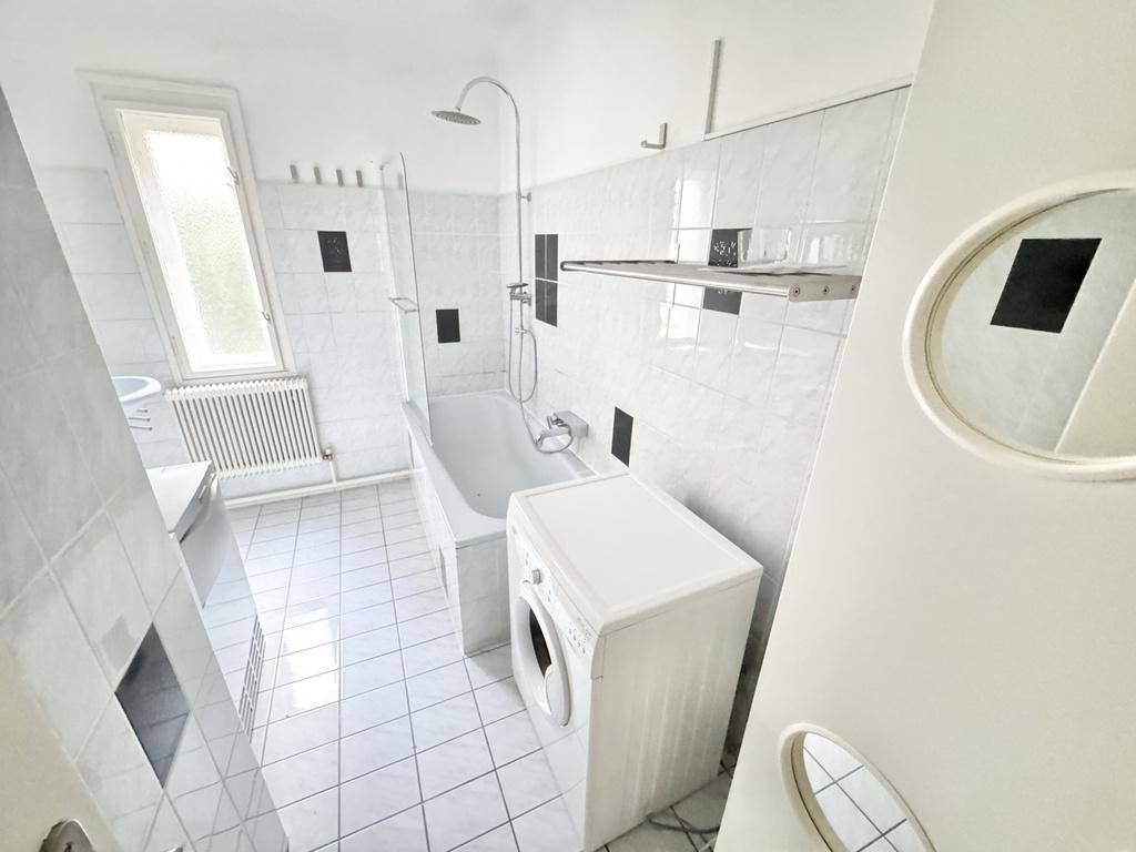 Helles Badezimmer mit weißen Fliesen, Badewanne, Dusche und einer Waschmaschine.