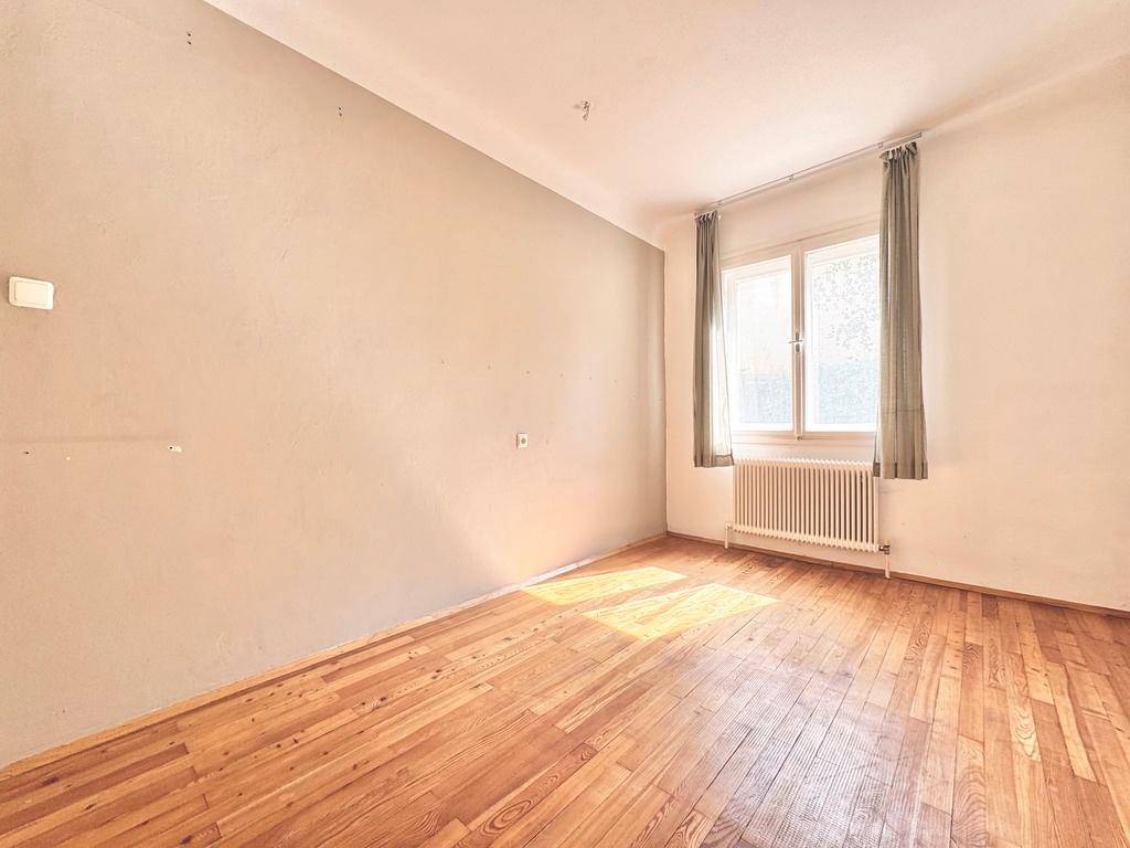 Sonniger Raum mit Holzboden, einer dezenten grauen Wand und einem Fenster mit Vorhängen.