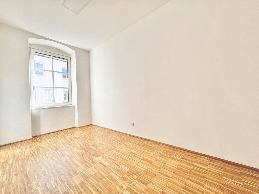 Helles Zimmer mit Parkettboden und einem Fenster, das viel Tageslicht hereinlässt.