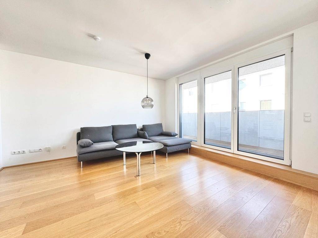 Großzügiger Wohnbereich mit hellem Holzboden, modernem Sofa und Zugang zu einem sonnigen Balkon.