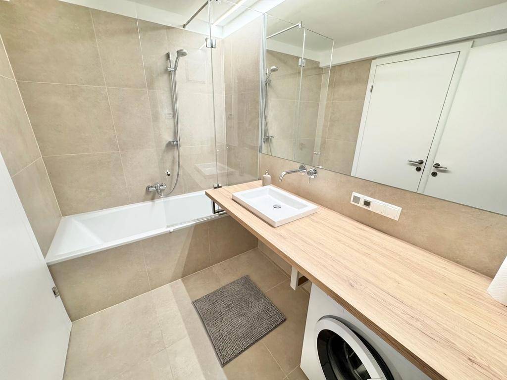 Modernes Badezimmer mit beigen Fliesen, Badewanne mit Duschfunktion und Waschbecken auf Holzplatte.
