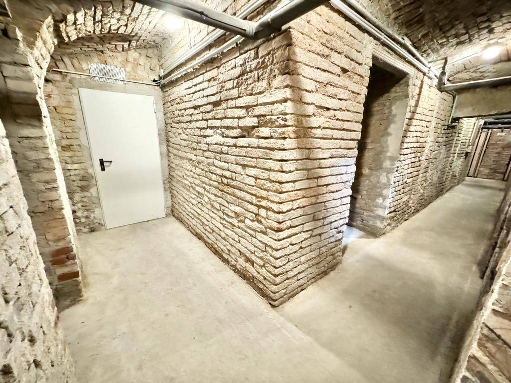 Historischer Kellerflur mit unverputzten Ziegelwänden, Betonboden und einer weißen Tür.