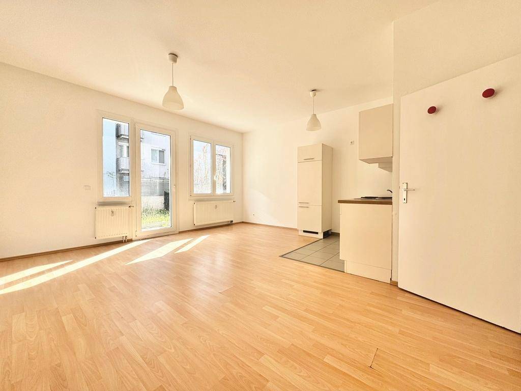 Heller Wohnbereich mit Laminatboden, großen Fenstern und einer integrierten Küchenzeile im Hintergrund.