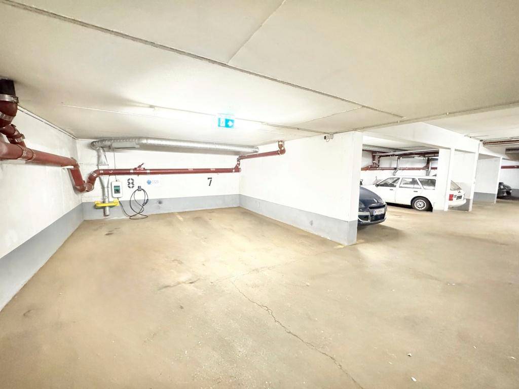 Tiefgaragenstellplatz mit Betonboden, sichtbaren Rohren und einem geparkten Fahrzeug im Hintergrund.