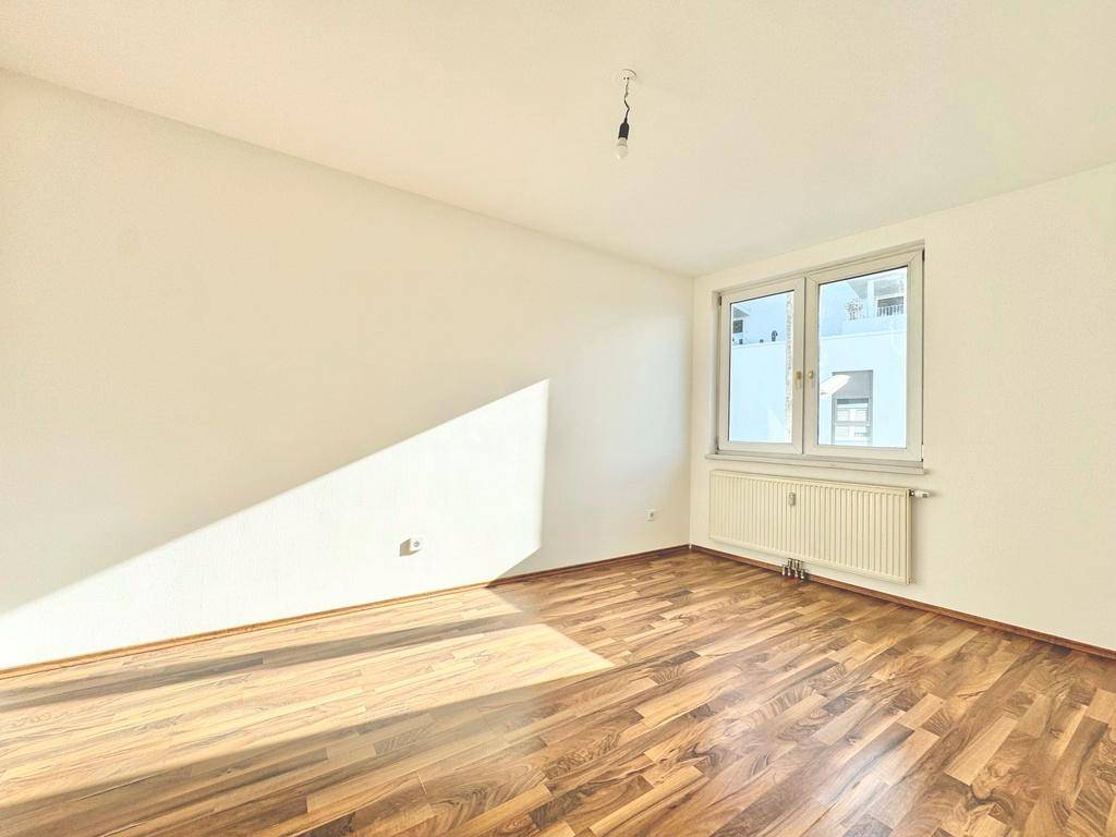 Leeres Zimmer mit Laminatboden und einem Fenster, das viel Sonnenlicht in den Raum lässt.