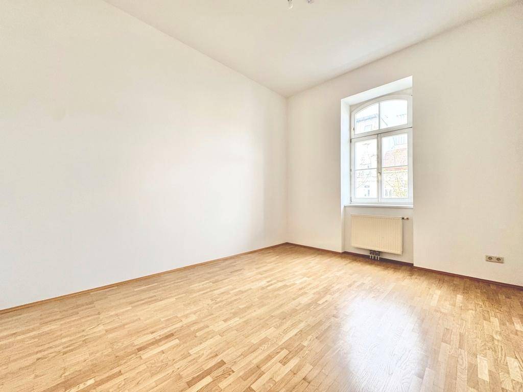 Gemütliches, leeres Schlafzimmer mit Parkettboden und einem großen Fenster, das viel Licht spendet.
