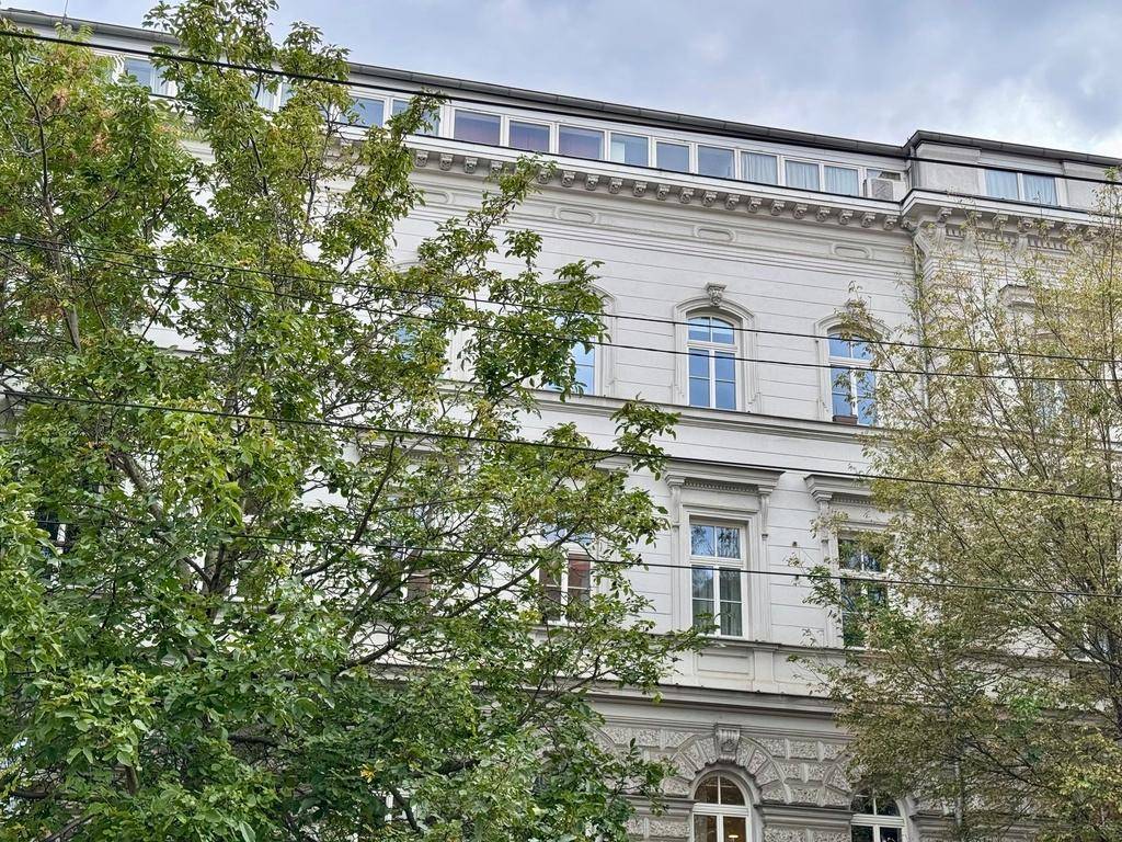 Historische Fassade eines Gebäudes mit verzierten Fenstern und klassischer Architektur, teilweise von Bäumen verdeckt.