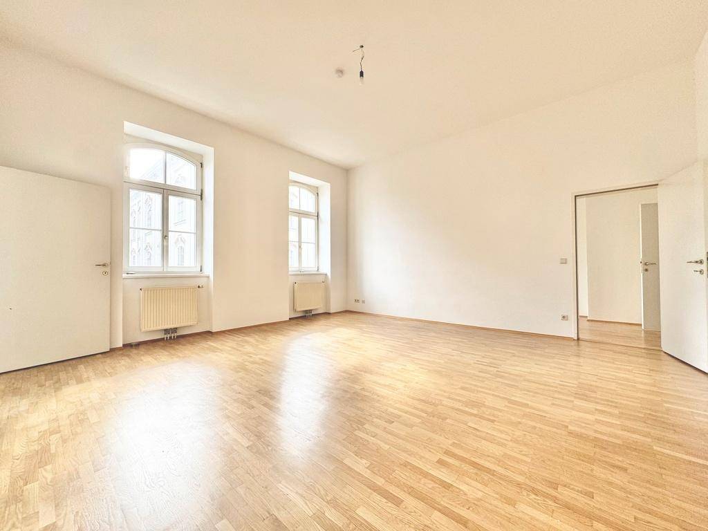 Geräumiges Zimmer mit hellem Parkettboden und großen Fenstern, die viel Tageslicht hereinlassen.