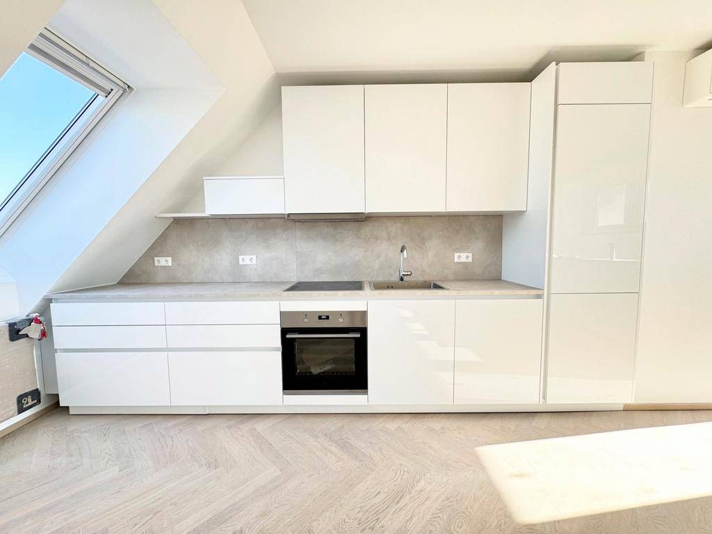 Moderne weiße Einbauküche mit glänzenden Fronten, integrierten Geräten und Arbeitsplatte unter einem Dachfenster.
