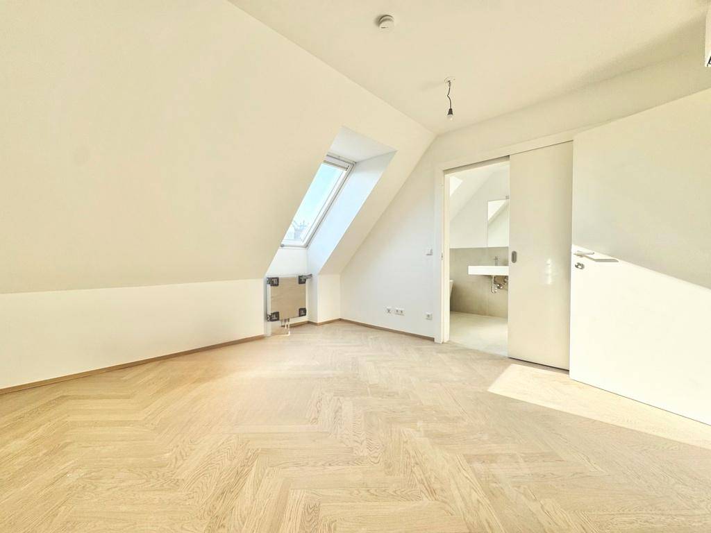Helles Dachzimmer mit Fischgrätparkett, Dachfenster und Zugang zum Badezimmer.
