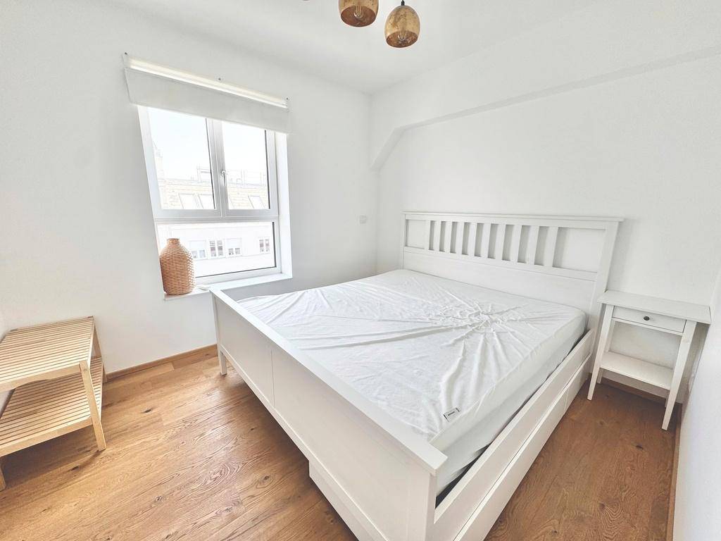 Gemütliches Schlafzimmer mit weißem Holzbett, Nachttisch und Holzboden, hell durch das Fenster.