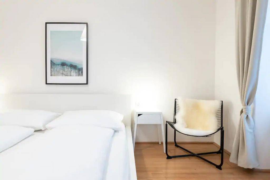 Schlafzimmer mit weißem Bett, modernem Stuhl und Vorhängen am Fenster.