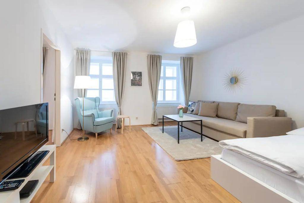 Geräumiges Wohnzimmer mit Holzboden, zwei Fenstern, einem Sofa und einem bequemen Sessel.