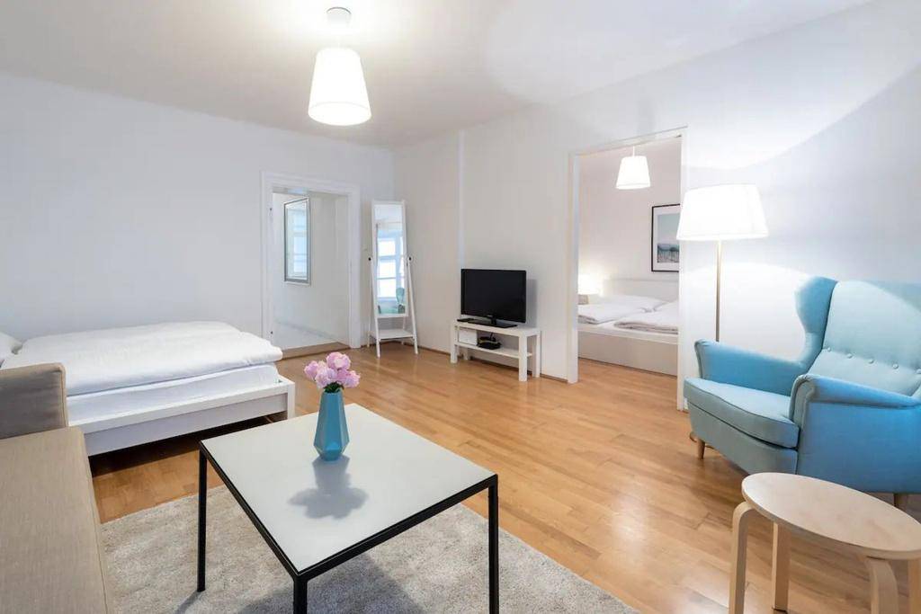 Helles Wohnzimmer mit Holzboden, einem blauen Sessel und Zugang zu einem weiteren Schlafbereich.