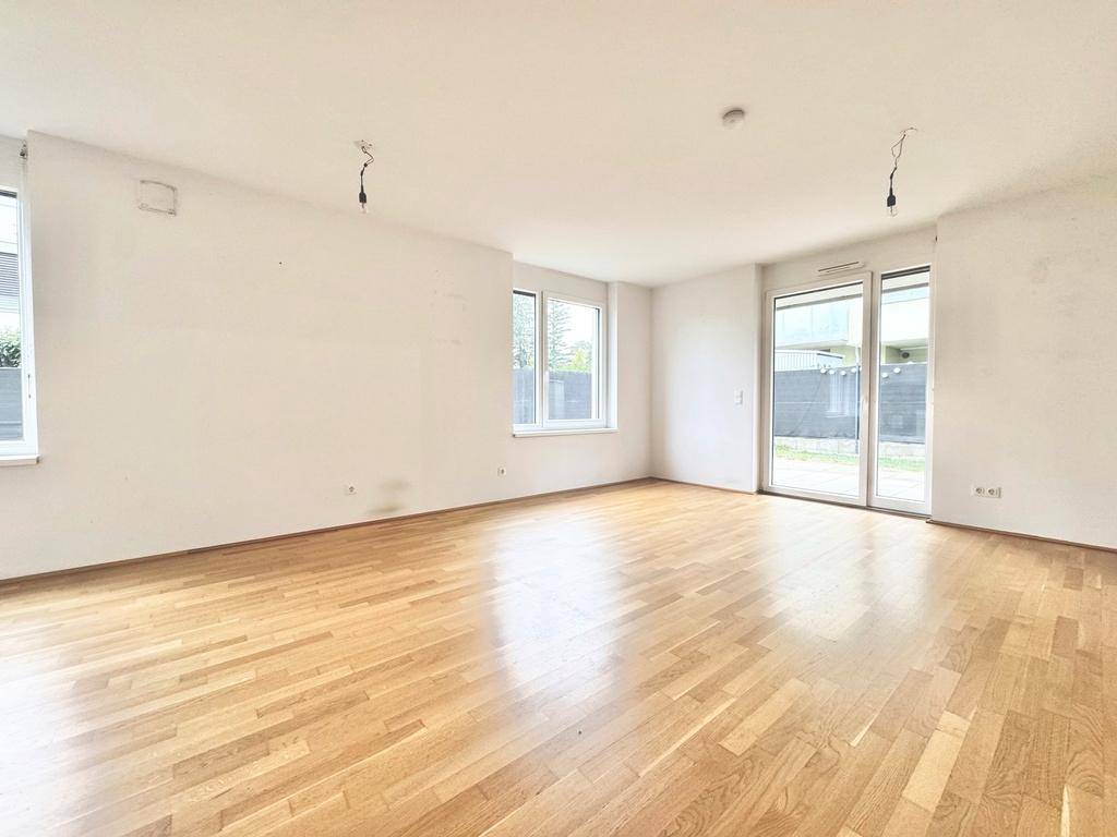 Helles, leeres Wohnzimmer mit Parkettboden und großzügigen Fenstern zum Außenbereich.