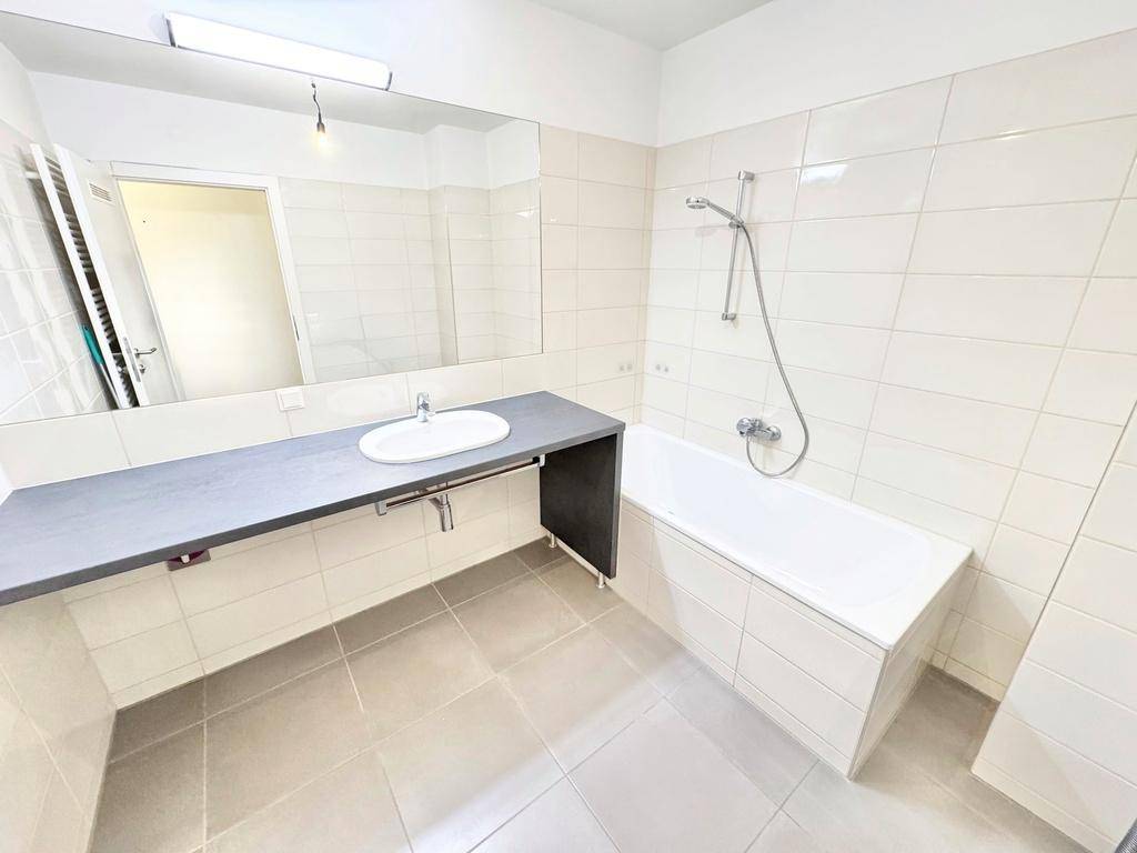 Funktionales Badezimmer mit Badewanne, Waschbecken und großen weißen Wand- und Bodenfliesen.