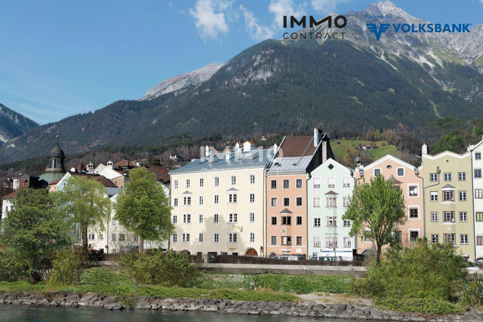 Historische Gebäude entlang eines Flusses mit majestätischem Bergblick im Hintergrund unter blauem Himmel.