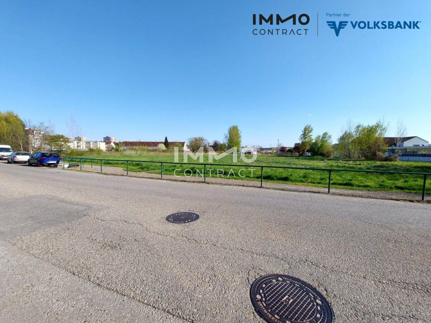 Blick auf eine Straße mit parkenden Autos, einem grünen Zaun und einer weiten Wiese im Hintergrund.