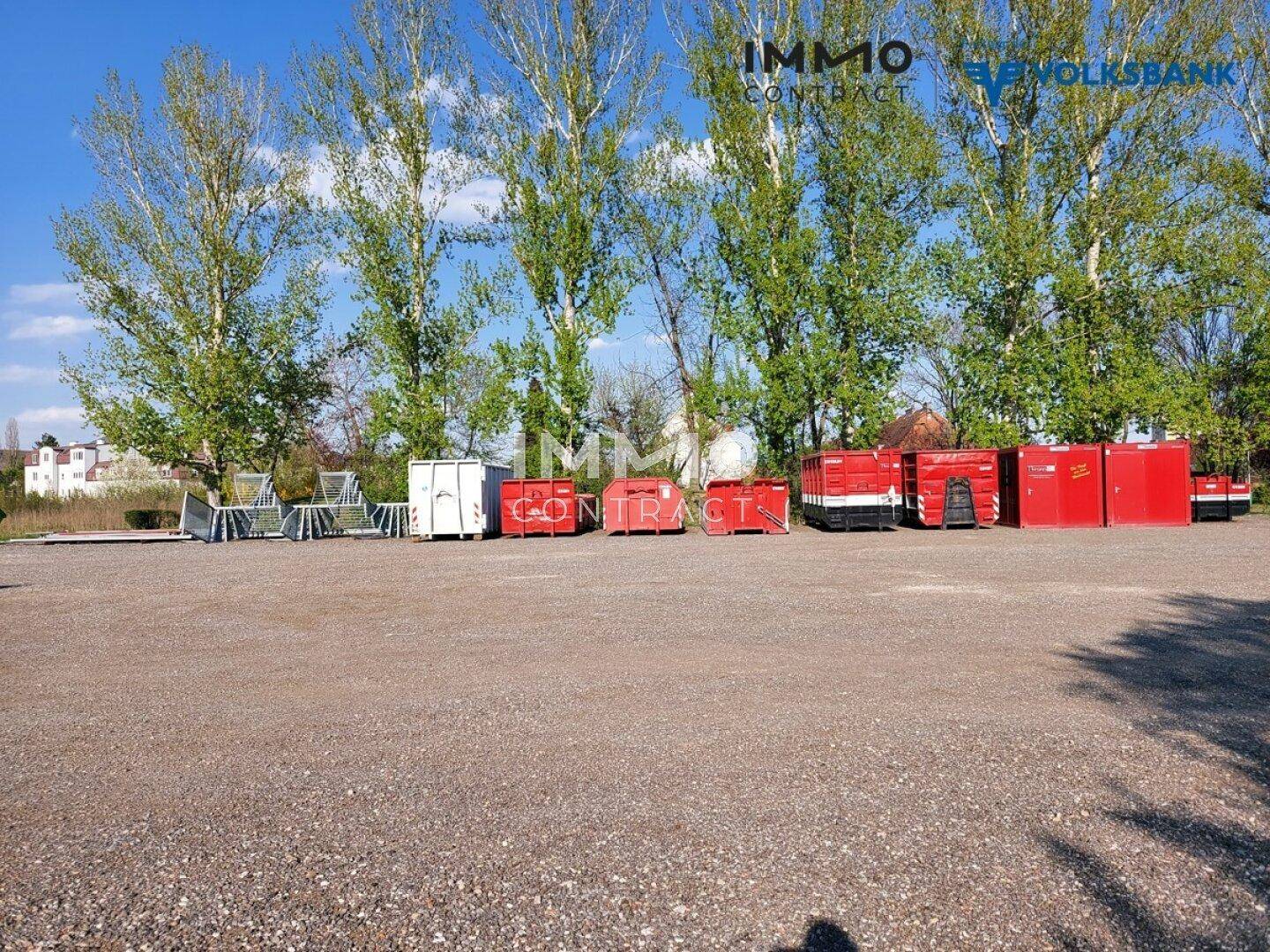 Unbefestigtes Grundstück mit roten Lagercontainern und Metallstrukturen unter hohen Bäumen.