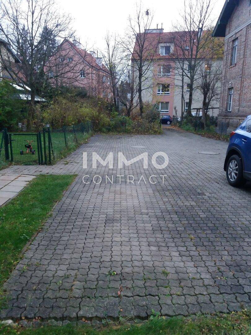 Gepflasterter Außenparkplatz mit umliegenden Bäumen und Gebäuden, umzäuntem Bereich.