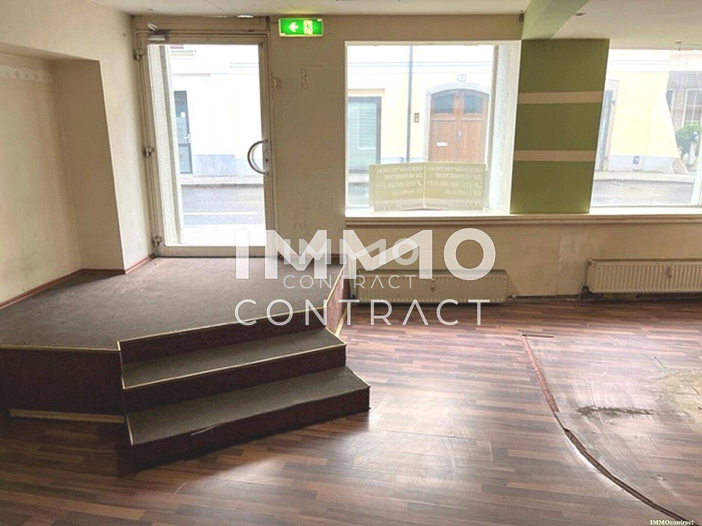 Innenansicht eines Ladenlokals mit Laminatboden, großer Fensterfront und einer erhöhten Plattform, die renovierungsbedürftig ist.