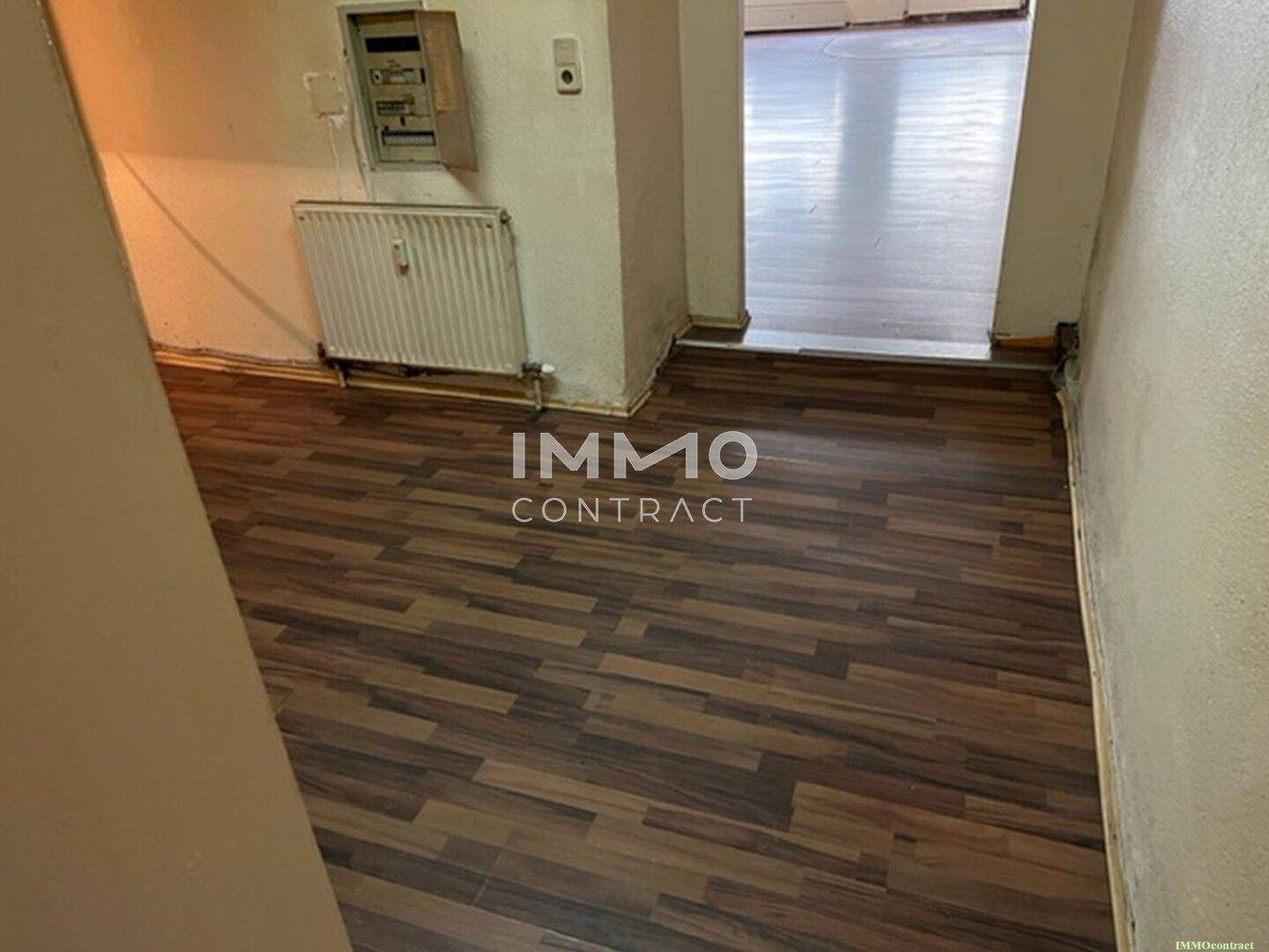 Flur mit dunklem Laminatboden und einem Heizkörper, der zu weiteren Räumen führt und eine einfache Ausstattung aufweist.