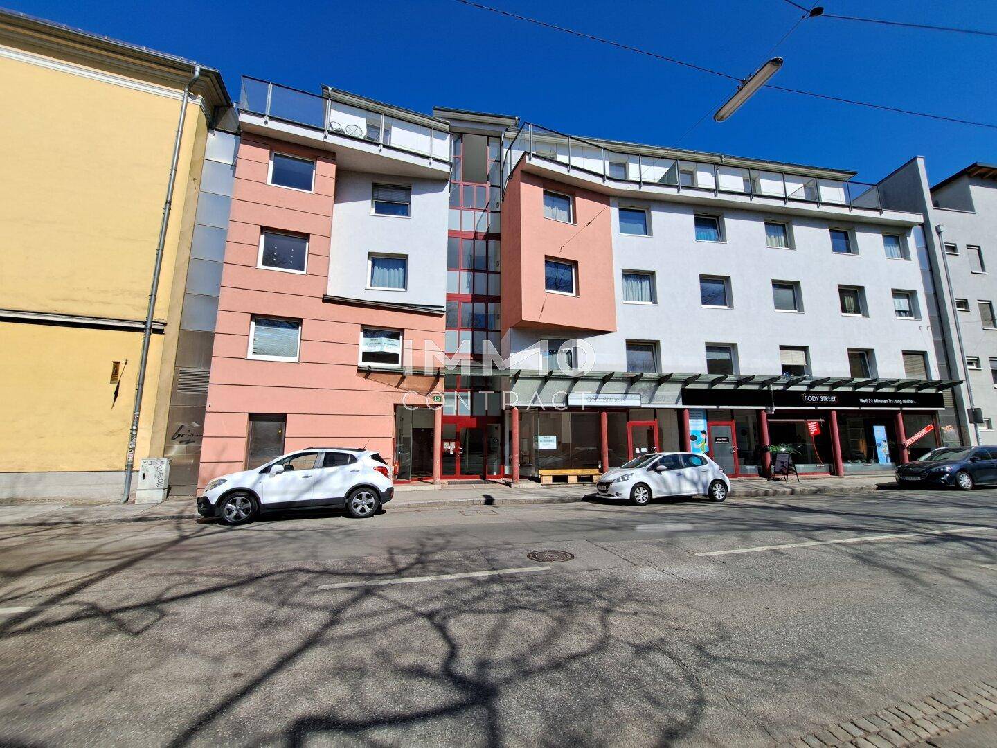 Mehrstöckiges Gebäude mit Geschäftsflächen im Erdgeschoss und Wohnungen darüber, an einer Straße gelegen.