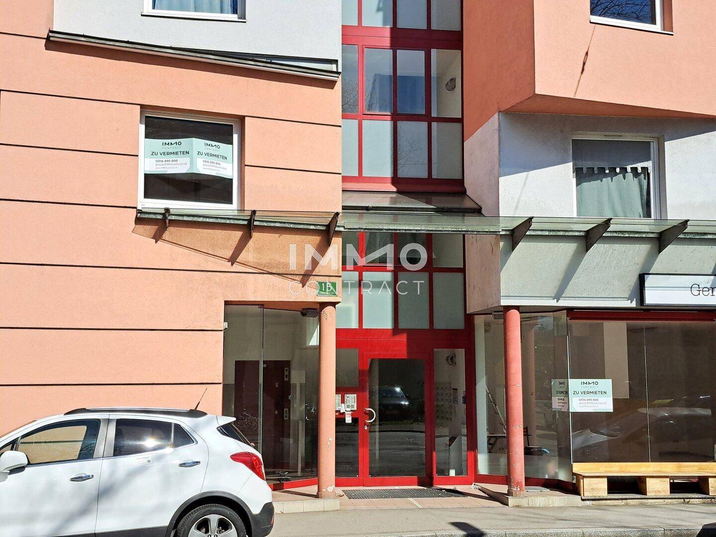 Eingangsbereich eines Gebäudes mit roten Türen und Fenstern, sowie Vermietungsschildern an der Fassade.