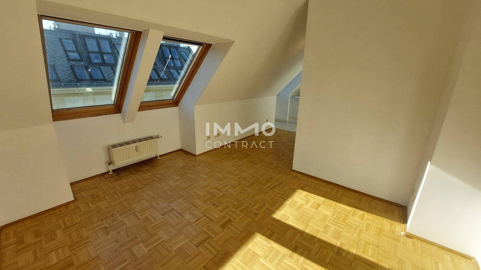 Helles Dachzimmer mit Parkettboden, zwei großen Dachfenstern und schrägen Wänden.