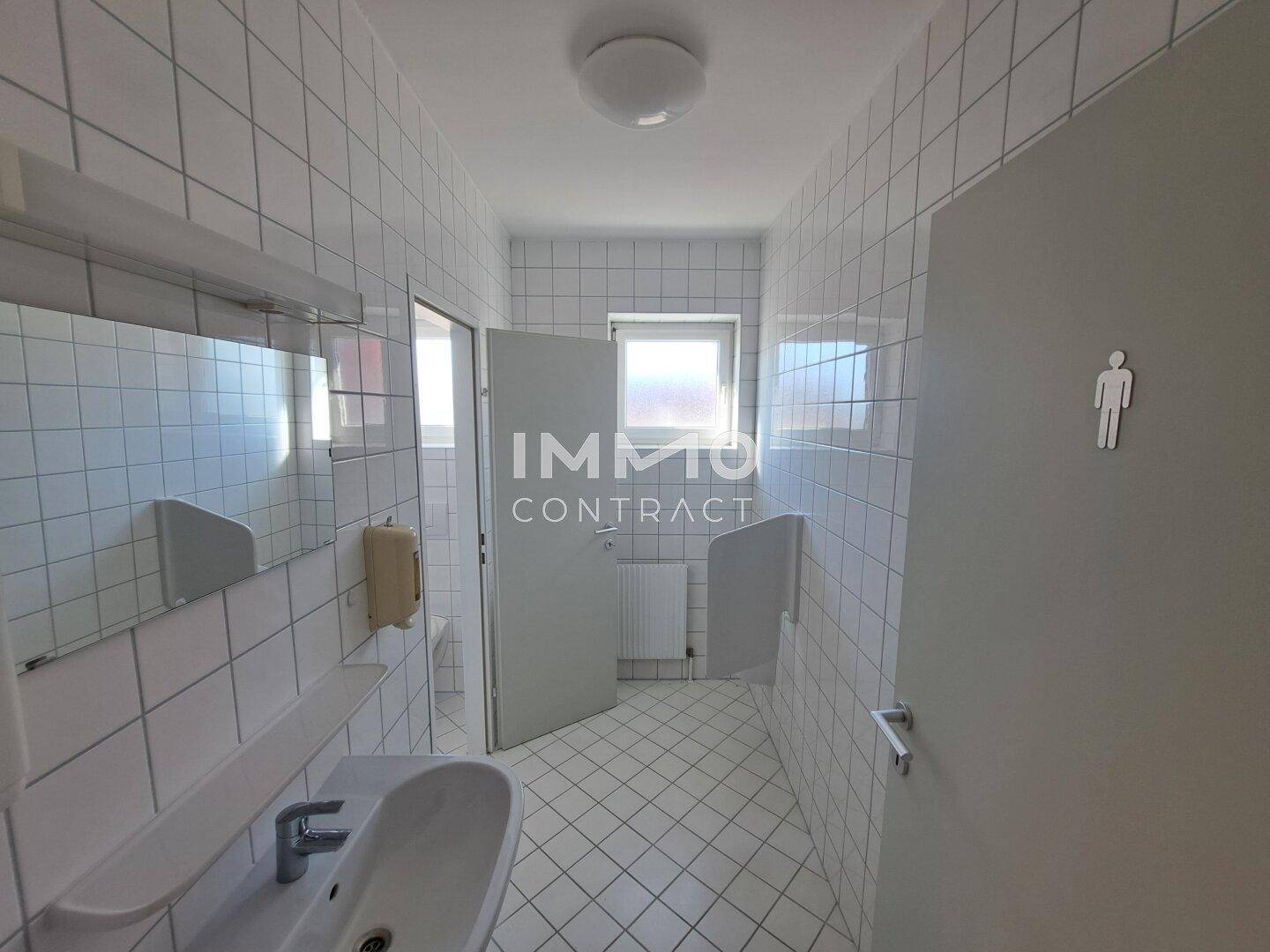 Geräumige Toilette mit weißen Fliesen, Urinal und Fenster, gut beleuchtet und gepflegt.