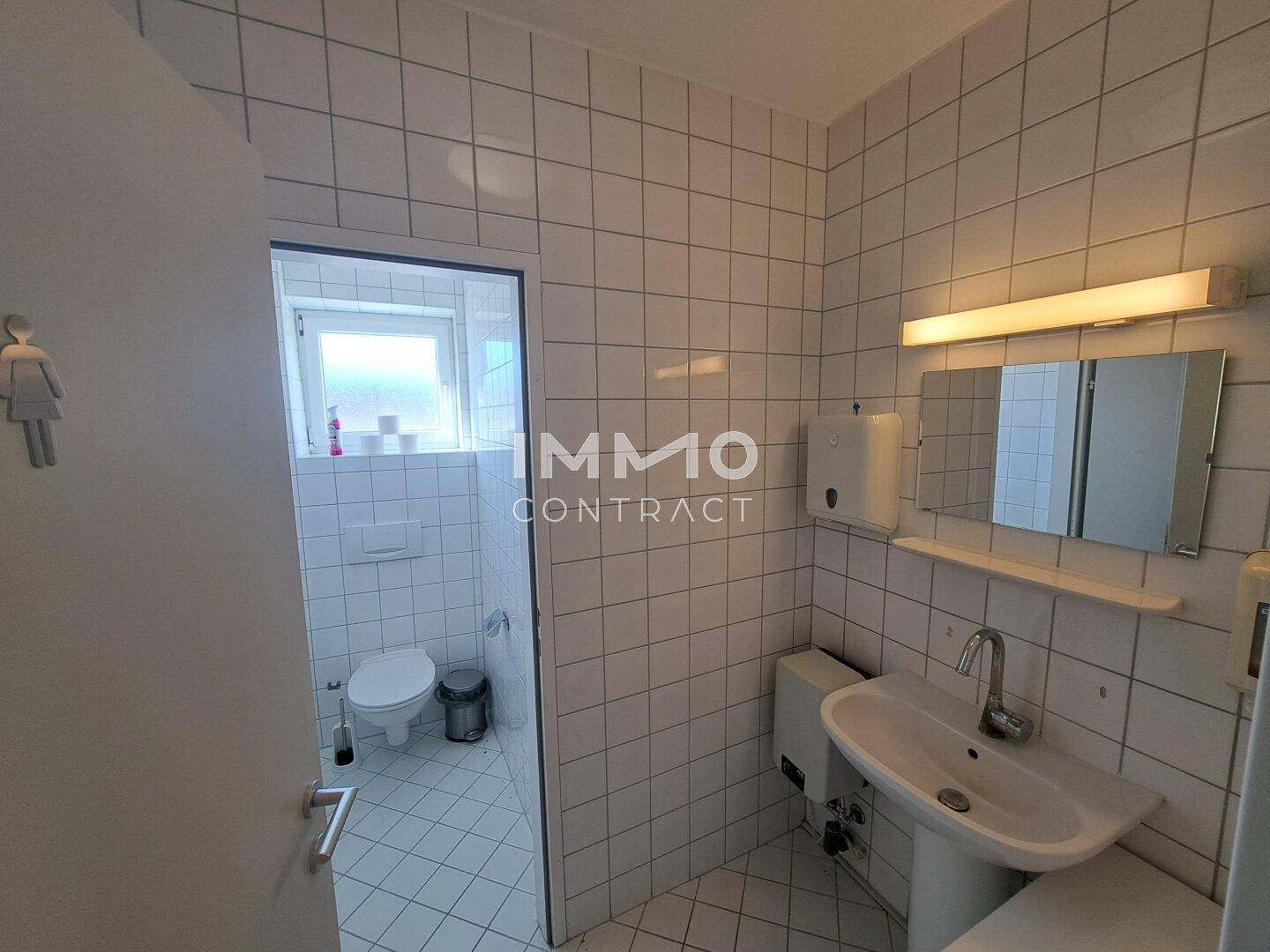 Helle Toilette mit weißen Fliesen, Waschbecken und Fenster, in gutem und sauberem Zustand.