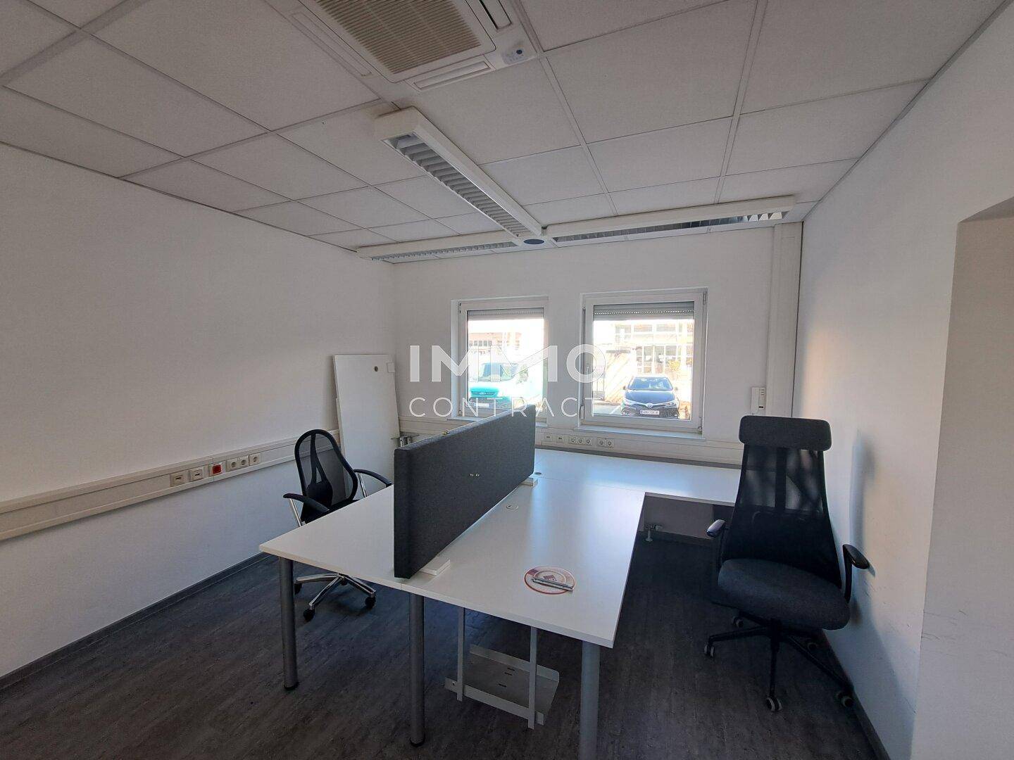 Modernes Büro mit zwei Arbeitsplätzen, großen Fenstern und abgehängter Decke mit Beleuchtung.