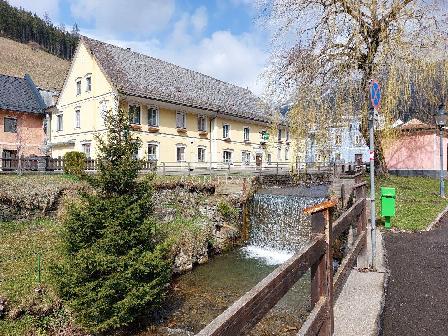 Idyllische Umgebung mit Bachlauf, Brücke und traditionellen Gebäuden vor einer Bergkulisse.