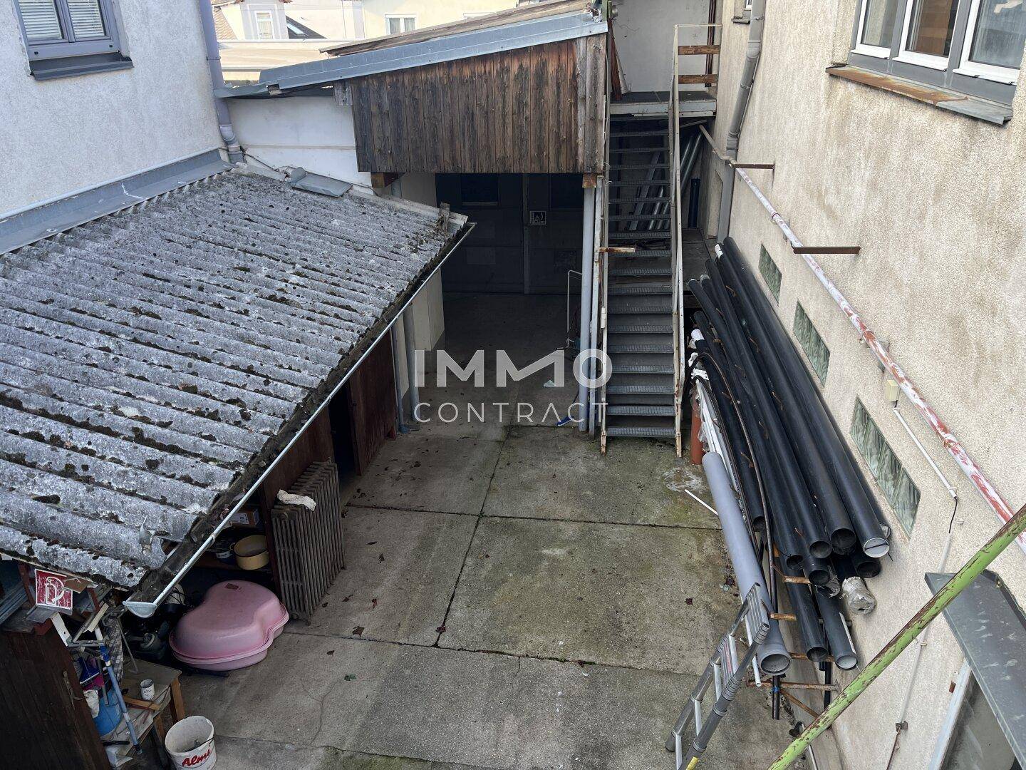 Hinterhofbereich mit Betonboden, einer Außentreppe und gelagerten Rohren.