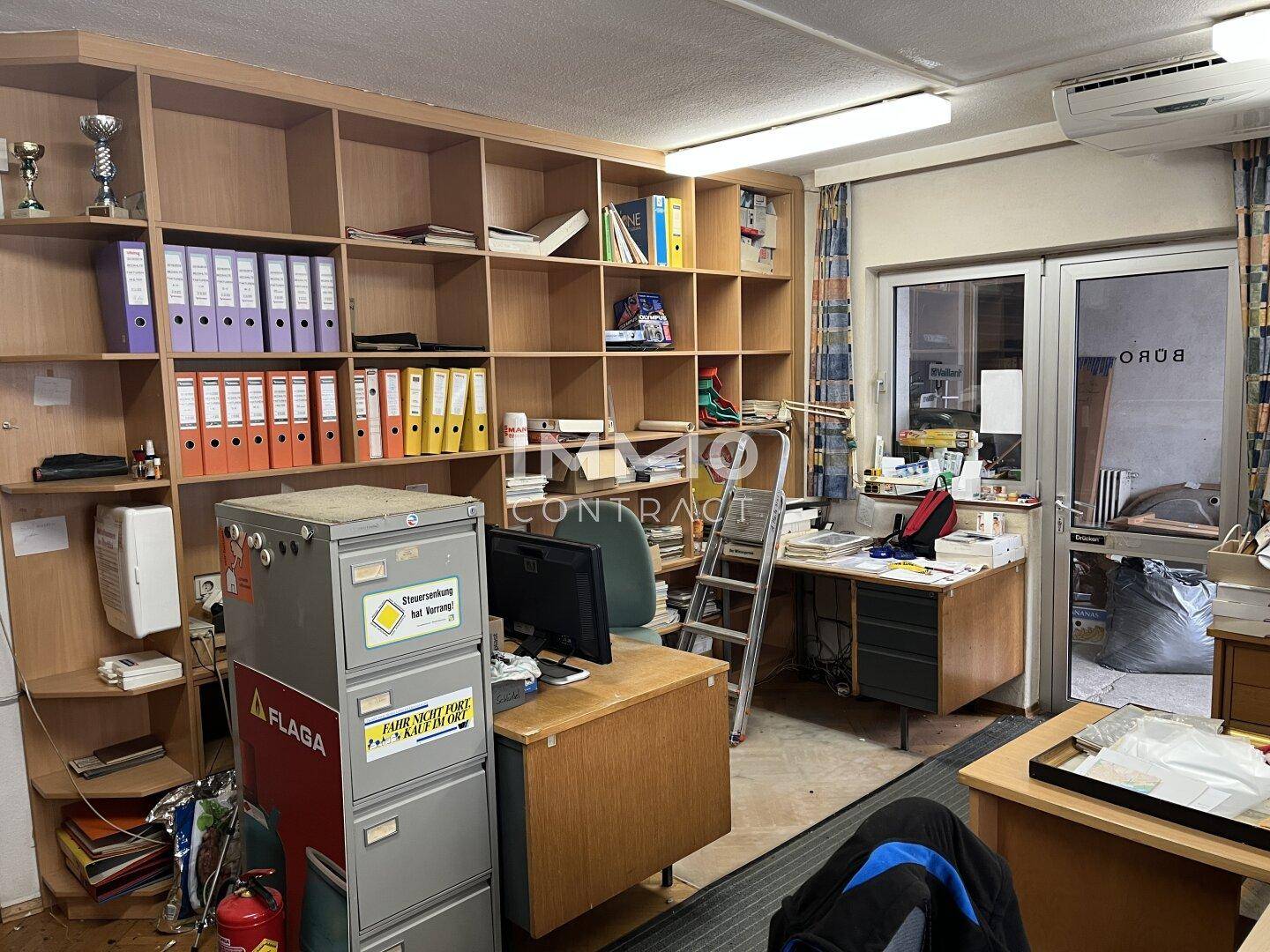 Unaufgeräumter Büroraum mit Regalen, Schreibtischen und einem Fenster mit Blick nach draußen.